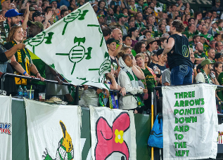 Photo of Portland Timbers fans holding flags depicting three arrows and written description.